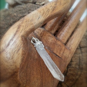 May include: A clear quartz crystal pendant with a silver-colored bail. The crystal is elongated and faceted, with a pointed tip. The pendant is displayed on a wooden surface.