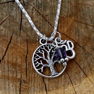 May include: Silver necklace with a tree of life pendant. The pendant has a detailed tree design within a circle. A purple gemstone and a charm with the number 30 are also attached. The necklace is displayed on a wooden surface.