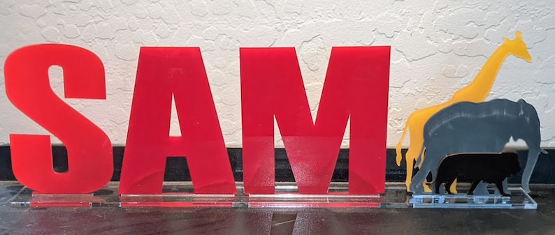 May include: Red acrylic letters spelling out the name "SAM" with a yellow giraffe and black elephant silhouettes behind the letters.