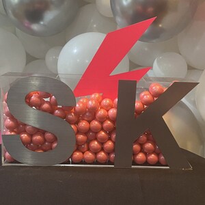 May include: A clear acrylic box filled with pink gumballs, with the letters "SK" in silver metal and a large pink lightning bolt cutout on top.