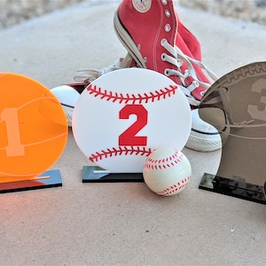 May include: Three sports-themed table numbers, a basketball with the number 1, a baseball with the number 2, and a football with the number 3. Each number is on a black stand and there is a small ball in front of each stand.