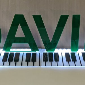 May include: Green acrylic letters spelling "DAVID" with a treble clef and a musical note on either side. The letters are illuminated from behind and are sitting on a white and black piano keyboard.