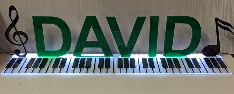 May include: Green acrylic letters spelling "DAVID" with a treble clef and a musical note on either side. The letters are illuminated from behind and are sitting on a white and black piano keyboard.