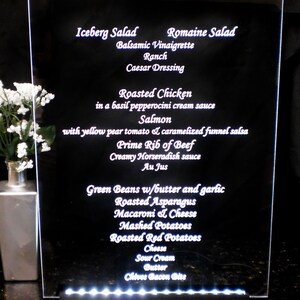 May include: A white illuminated menu board with a black background listing various dinner options, including salads, roasted chicken, salmon, prime rib of beef, green beans, roasted asparagus, macaroni and cheese, mashed potatoes, roasted red potatoes, cheese, sour cream, butter, and chives bacon bits.