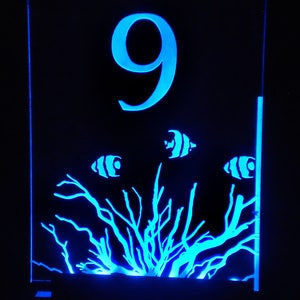 May include: A blue acrylic table number with the number 9 and a design of coral and fish. The number is illuminated from behind.