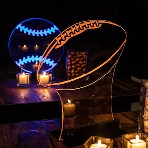 May include: An illuminated football-shaped acrylic lamp with an orange glow, standing on a black base. A baseball-shaped lamp with blue illumination is in the background. Small candles in glass holders are scattered around the lamps.