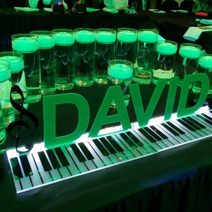 May include: A green illuminated piano keyboard with the name "DAVID" spelled out in large green letters. There are several clear glass cylinders filled with green liquid and a black treble clef on the table.
