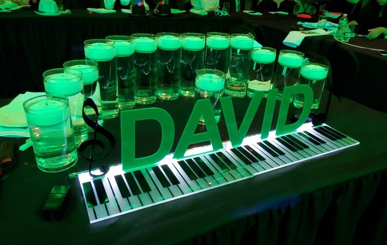 May include: A green illuminated piano keyboard with the name "DAVID" spelled out in large green letters. There are several clear glass cylinders filled with green liquid and a black treble clef on the table.