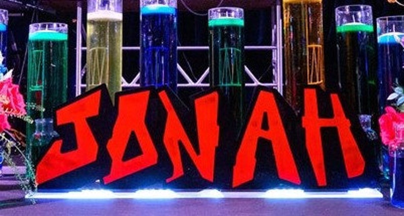 May include: A red and black sign with the name "JONAH" in large, bold letters. The sign is decorated with colourful glass cylinders filled with liquids.