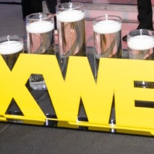 May include: Yellow letters spelling out the name "MAXWELL" with clear glass cylinders filled with water and floating candles in front of the letters.