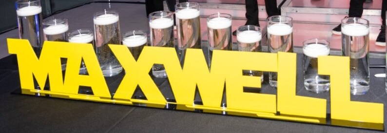 May include: Yellow letters spelling out the name "MAXWELL" with clear glass cylinders filled with water and floating candles in front of the letters.