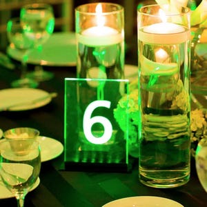 May include: A green acrylic table number with the number "6" in white. The table number is on a table with green and white floral arrangements and two clear glass cylinders with candles.