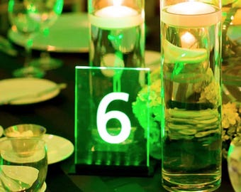 May include: A green acrylic table number with the number "6" in white. The table number is on a table with green and white floral arrangements and two clear glass cylinders with candles.