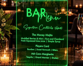 May include: A rectangular, transparent acrylic bar menu sign with bright green LED illumination. The text reads: BAR Menu, Signature Cocktails (Adult), The Money Mojito, Muddled Berries & Mint | Rum and Chambord, Fresh Squeezed Lime Juice | Simple Syrup, Players Card, Bourbon | Sweet Vermouth | Bitters, Ash Conn Margherita, Tequila | Grand Marnier | Shaken.