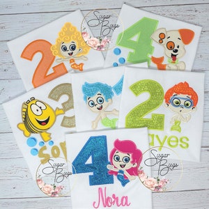 May include: Five white t-shirts with colorful embroidered numbers and characters from the Bubble Guppies cartoon. The numbers are 2, 3, 4, 2, and 4. The characters include Molly, Gil, Goby, and Oona. The shirts are for a birthday party.