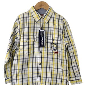 May include: Yellow, white, and black plaid button-down shirt with two chest pockets. The shirt has a collar and long sleeves. A tag with the word "POINTER" is attached. A patch with the text "POINTER CAMPUS CLASS OF 1979" is on the shirt.