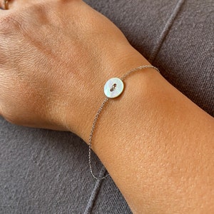 Può includere: Un delicato braccialetto a catena d'argento con un ciondolo a forma di bottone in madreperla bianca.