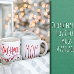 May include: Two white ceramic mugs with red lettering. One mug says "North Pole Hot Chocolate" and the other mug says "Mom". The mugs are sitting on a white and gray blanket with a blurred background of Christmas lights.