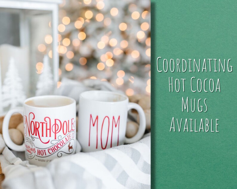 May include: Two white ceramic mugs with red lettering. One mug says "North Pole Hot Chocolate" and the other mug says "Mom". The mugs are sitting on a white and gray blanket with a blurred background of Christmas lights.