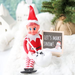 May include: A toy elf with a red suit, white collar, and red and white striped legs holding a small black shovel. The elf sits next to a miniature wooden easel displaying a sign that reads LET'S MAKE SNOW! with a graphic of a snowman.