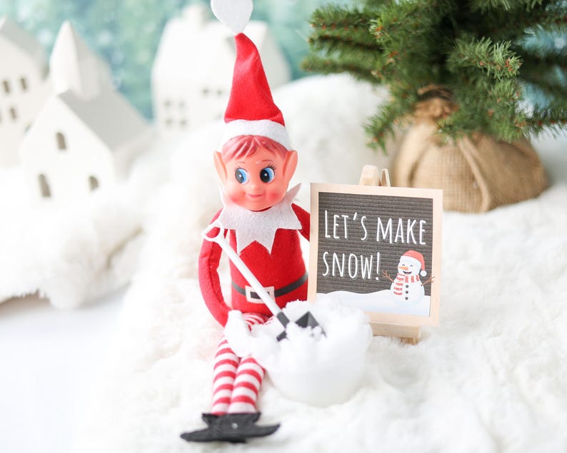 May include: A toy elf with a red suit, white collar, and red and white striped legs holding a small black shovel. The elf sits next to a miniature wooden easel displaying a sign that reads LET'S MAKE SNOW! with a graphic of a snowman.
