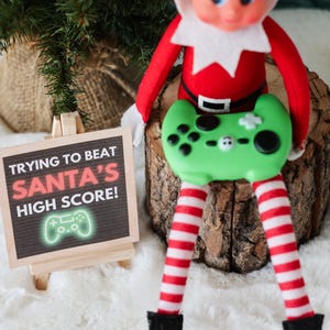 May include: A Christmas elf doll wearing a red and white outfit, holding a green video game controller. A small chalkboard sign reads "Trying to beat Santa's high score!" with a glowing green game controller icon. The scene is set on a white, fluffy surface.