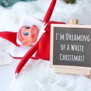 May include: A red and white elf doll lying down next to a small wooden easel holding a gray felt letter board. The board features white plastic letters that read, I'M DREAMING OF A WHITE CHRISTMAS! The elf has a pointed hat and blue eyes.