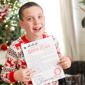 May include: A child wearing red and white Christmas pajamas holds a letter from Santa Claus. The letterhead reads "From the Desk of Santa Claus" and includes a personalized message and a Santa Claus signature.
