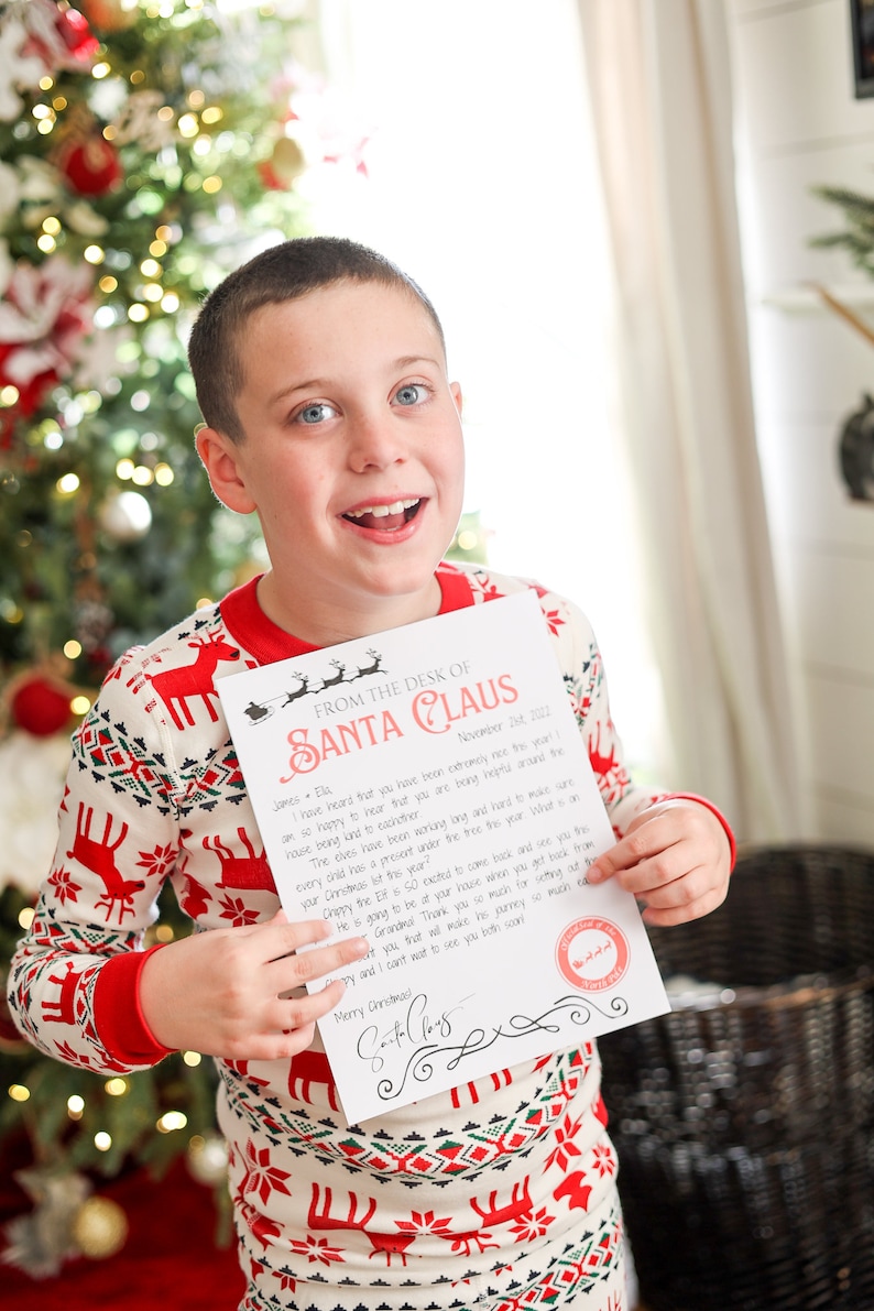 May include: A child wearing red and white Christmas pajamas holds a letter from Santa Claus. The letterhead reads "From the Desk of Santa Claus" and includes a personalized message and a Santa Claus signature.
