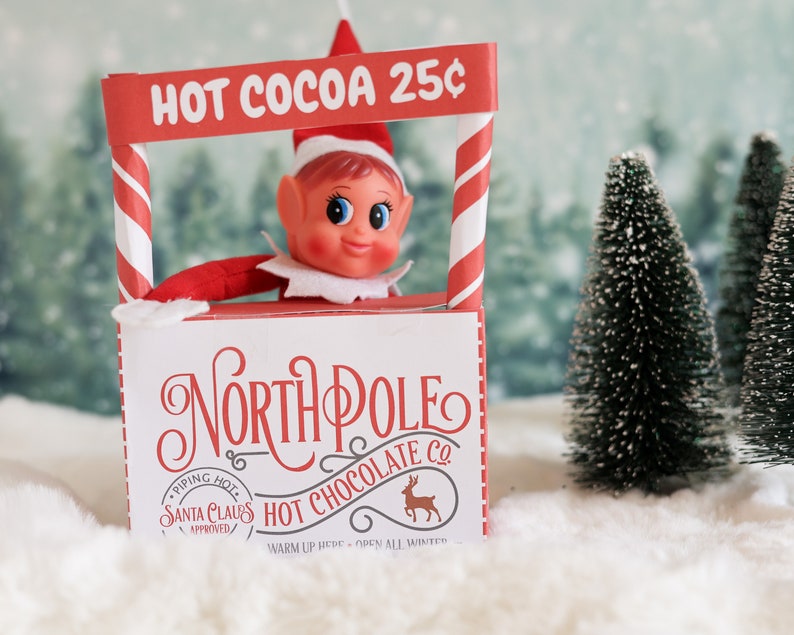 May include: A toy elf wearing a red and white striped hat stands behind a red and white striped counter with a sign that reads "HOT COCOA 25&cent;". The counter is decorated with a white sign that reads "NORTH POLE HOT CHOCOLATE CO. PIPING HOT SANTA CLAUS APPROVED WARM UP HERE OPEN ALL WINTER".