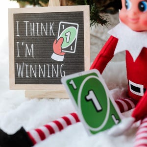 May include: A festive scene featuring an elf doll holding an Uno card with the number 1. A sign reads "I THINK I'M WINNING" with a graphic of a hand holding an Uno card. The elf is dressed in a red and white outfit.