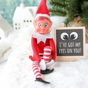 May include: A posable elf doll with a red suit, white collar, and red and white striped legs, wearing a red hat. Next to the doll is a small square letter board with a light wood frame, displaying two large googly eyes and the text I'VE GOT MY EYES ON YOU!