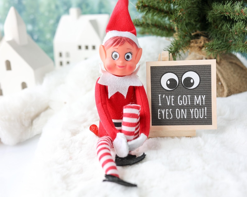May include: A posable elf doll with a red suit, white collar, and red and white striped legs, wearing a red hat. Next to the doll is a small square letter board with a light wood frame, displaying two large googly eyes and the text I'VE GOT MY EYES ON YOU!