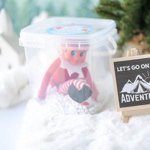 May include: A clear plastic container holding a small elf doll with red clothing and striped legs, sitting on white foam beads. Next to it is a small wooden easel displaying a black sign with white text that reads LET'S GO ON AN ADVENTURE.