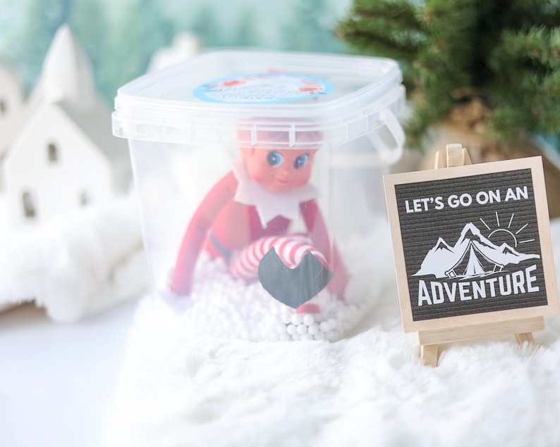 May include: A clear plastic container holding a small elf doll with red clothing and striped legs, sitting on white foam beads. Next to it is a small wooden easel displaying a black sign with white text that reads LET'S GO ON AN ADVENTURE.