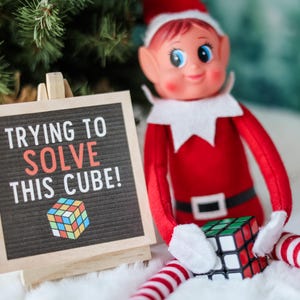 Op de afbeelding: Een elfpop in een rood pak en hoed houdt een Rubik's Cube vast. Een klein bordje leest "TRYING TO SOLVE THIS CUBE!" met een kleurrijke kubusillustratie. De scène speelt zich af tegen een achtergrond van een kerstboom en witte kunstsneeuw.