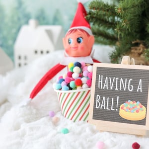 May include: A festive elf doll with a red hat and suit sits behind a paper cup filled with colorful miniature pom-poms. Next to the cup is a small wooden easel holding a gray felt letter board with the white text HAVING A BALL! and a graphic.