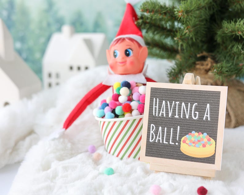 May include: A festive elf doll with a red hat and suit sits behind a paper cup filled with colorful miniature pom-poms. Next to the cup is a small wooden easel holding a gray felt letter board with the white text HAVING A BALL! and a graphic.