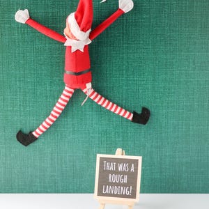 May include: A plush elf doll in a red suit with white trim, a pointed hat, and red-and-white striped legs, positioned as if stuck to a wall. A speech bubble says OUCH! A small wooden easel holds a black sign with the text THAT WAS A ROUGH LANDING!