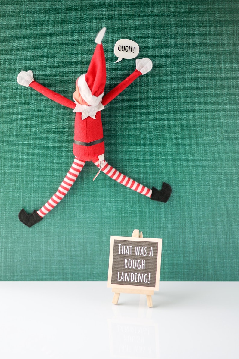 May include: A plush elf doll in a red suit with white trim, a pointed hat, and red-and-white striped legs, positioned as if stuck to a wall. A speech bubble says OUCH! A small wooden easel holds a black sign with the text THAT WAS A ROUGH LANDING!