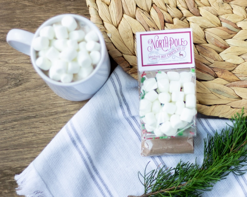 Op de afbeelding: Een witte mok gevuld met mini marshmallows, een klein zakje North Pole Hot Chocolate mix met mini marshmallows, een wit-blauwe gestreepte theedoek en een groene dennentak op een houten oppervlak.