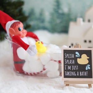 May include: A Christmas elf doll taking a bath in a white tub with a yellow rubber ducky. A sign reads "Splish Splash I'm Just Taking A Bath!"