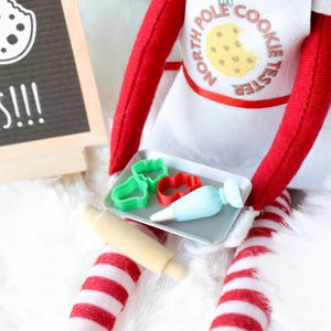 May include: A red and white striped doll wearing a white apron with the text NORTH POLE COOKIE TESTER and a cookie graphic. The doll holds a silver tray with two small cookie cutters, a light blue piping bag, and a wooden rolling pin.