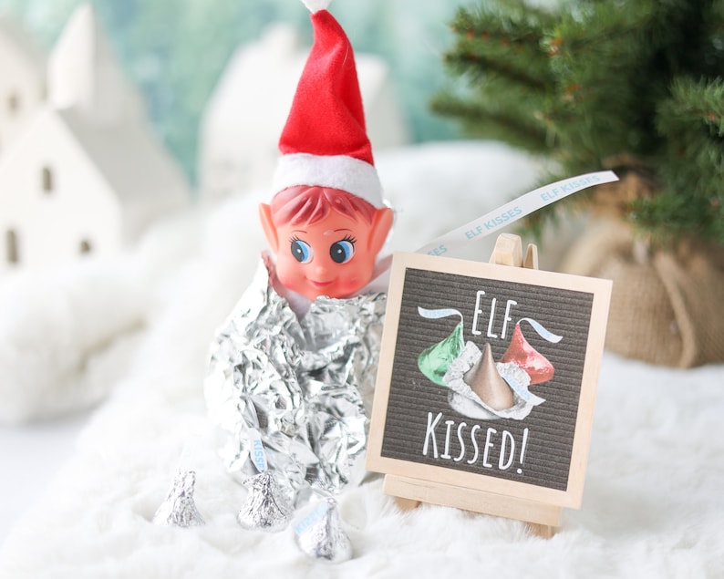 May include: A plastic elf doll wearing a red and white hat, wrapped in crinkled aluminum foil, positioned next to a small wooden easel holding a black sign with the white text ELF KISSED and illustrations of three chocolate candy drops.