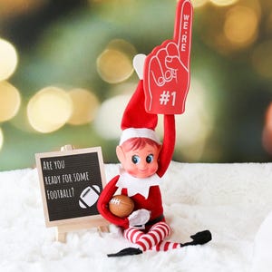 May include: A red and white elf doll holding a football and a foam finger that says "We're #1". The elf is sitting on a white fluffy surface with a chalkboard sign that says "Are you ready for some football?"