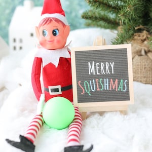 May include: A red and white elf doll with a pointed hat, wearing a black belt with a white buckle, sits next to a small wooden easel holding a black letter board that displays the white and colorful text Merry SQUISHMAS and a lime green ball.