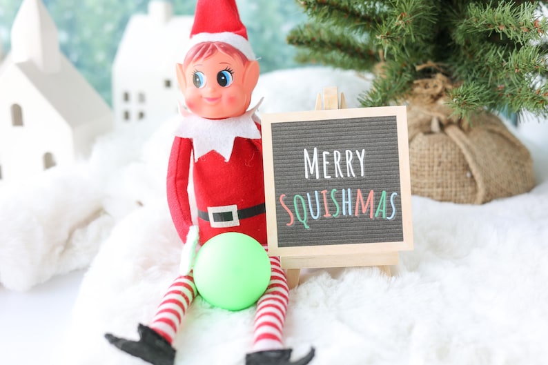 May include: A red and white elf doll with a pointed hat, wearing a black belt with a white buckle, sits next to a small wooden easel holding a black letter board that displays the white and colorful text Merry SQUISHMAS and a lime green ball.