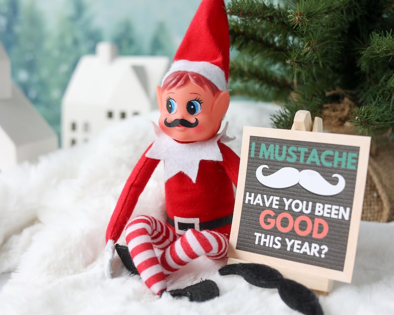 May include: A red and white elf doll with a black mustache, wearing a pointed hat and striped leggings. Next to the doll is a small wooden easel holding a sign that reads: I MUSTACHE HAVE YOU BEEN GOOD THIS YEAR? A black felt mustache lies below the sign.
