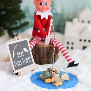 May include: An elf doll in a red suit and striped legs sits on a wooden stump, fishing with a red and white striped pole. A sign that says "Here Fishy Fishy!" is in front of the elf. A blue pond with rocks and fish crackers completes the scene.
