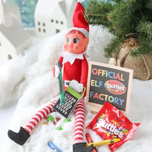 May include: A plush elf doll with a red suit and hat, red and white striped legs, and black boots. The elf holds a green crayon and a small metal grater. Next to it is a sign reading OFFICIAL ELF SKITTLES FACTORY and a bag of Skittles candy.